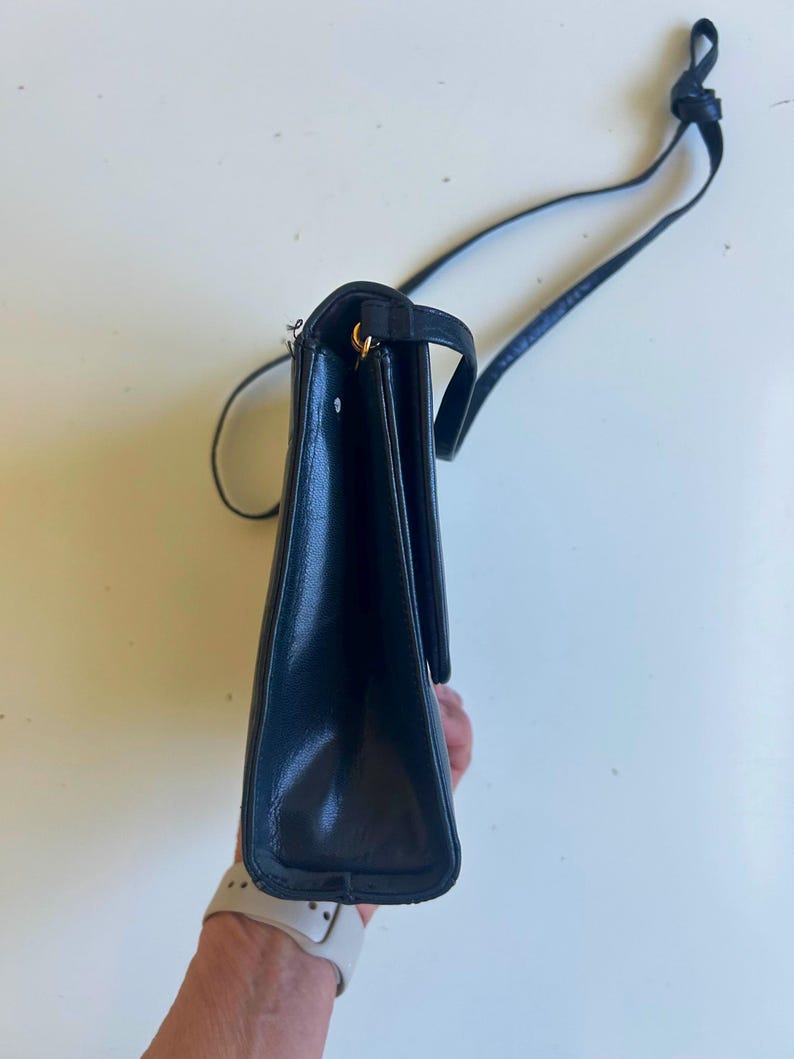 Pode incluir: Bolsa transversal de couro preta com uma al&ccedil;a longa. A bolsa tem formato retangular com fecho de aba e detalhes dourados. A al&ccedil;a &eacute; fina e ajust&aacute;vel. A bolsa &eacute; mostrada de lado.