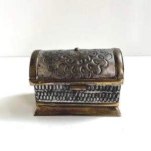 May include: A small rectangular chest with a hinged, arched lid. The exterior features a metallic silver finish with embossed floral patterns on the top and a textured, repeating vertical line design on the front, accented by brass-toned metal trim.