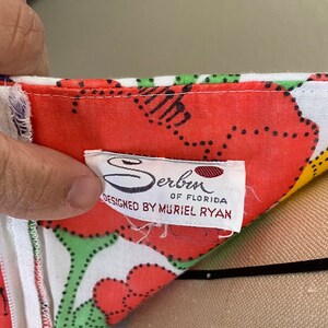 May include: Close-up of a colorful floral print fabric with a white zipper. The fabric features large red flowers with green and black accents on a white background. A tag reads "Serben of Florida, Designed by Muriel Ryan."
