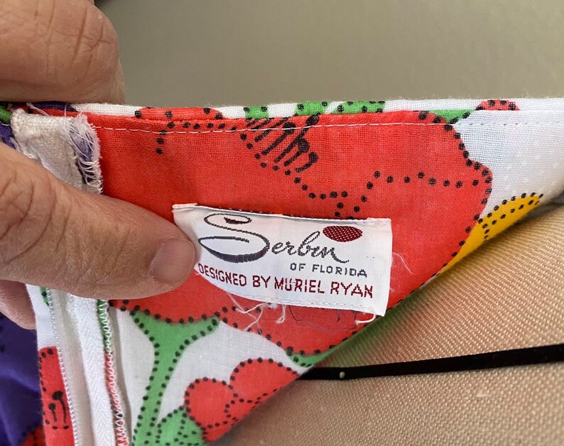May include: Close-up of a colorful floral print fabric with a white zipper. The fabric features large red flowers with green and black accents on a white background. A tag reads "Serben of Florida, Designed by Muriel Ryan."