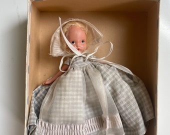 Puede incluir: Una muñeca vintage con cabello rubio y un vestido de cuadros azules y blancos. La muñeca lleva un gorro blanco y está sentada en una caja con fondo blanco.