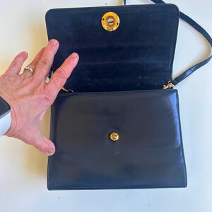 Pode incluir: Uma bolsa de ombro de couro preto com fecho de aba e ferragens douradas. A bolsa tem formato retangular e uma al&ccedil;a longa e fina. O interior &eacute; forrado com um tecido escuro. A bolsa est&aacute; aberta, revelando o interior.