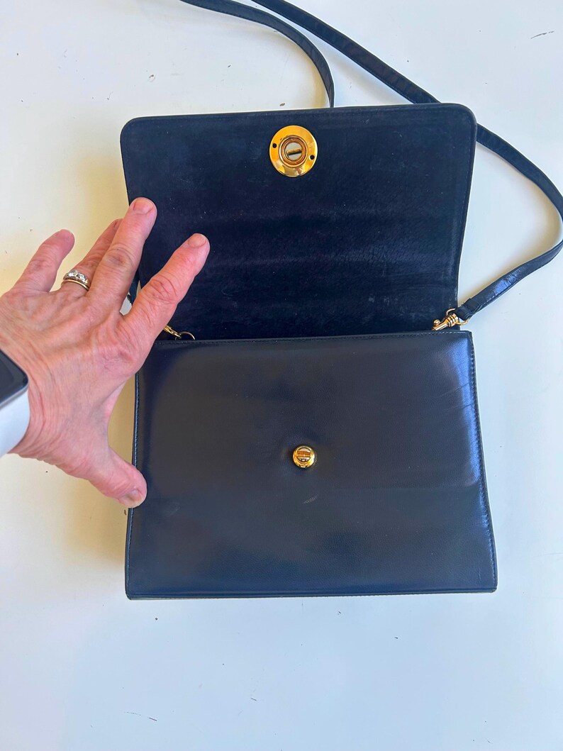 Pode incluir: Uma bolsa de ombro de couro preto com fecho de aba e ferragens douradas. A bolsa tem formato retangular e uma al&ccedil;a longa e fina. O interior &eacute; forrado com um tecido escuro. A bolsa est&aacute; aberta, revelando o interior.