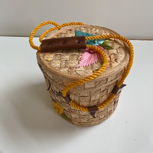 Op de afbeelding: Een ronde geweven strooien tas met een gele touwhandvat en bruine leren accenten. De tas is versierd met kleurrijke geborduurde bloemen.