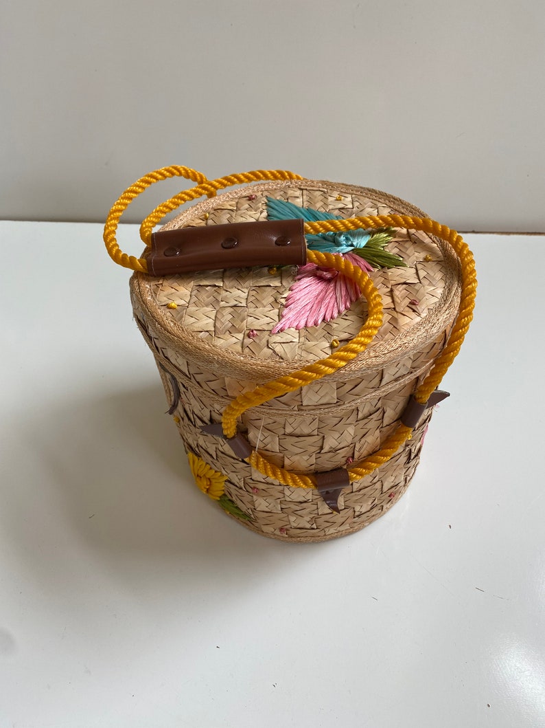 Op de afbeelding: Een ronde geweven strooien tas met een gele touwhandvat en bruine leren accenten. De tas is versierd met kleurrijke geborduurde bloemen.