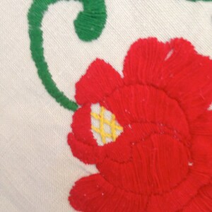 K&ouml;nnte beinhalten: Nahaufnahme einer roten gestickten Blume mit gelber Mitte und gr&uuml;nen Bl&auml;ttern auf wei&szlig;em Stoff.