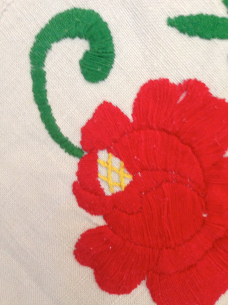 K&ouml;nnte beinhalten: Nahaufnahme einer roten gestickten Blume mit gelber Mitte und gr&uuml;nen Bl&auml;ttern auf wei&szlig;em Stoff.