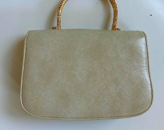 May include: A vintage handbag with a light beige exterior and a gold-colored, braided handle. The rectangular bag has rounded corners and a textured surface, suggesting a leather or leather-like material. The handle is arched and securely attached.