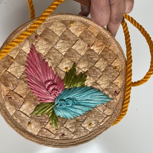 Op de afbeelding: Een ronde geweven strooien tas met een gele touwhandvat. De tas heeft een bloemenmotief met roze, groene en blauwe accenten.