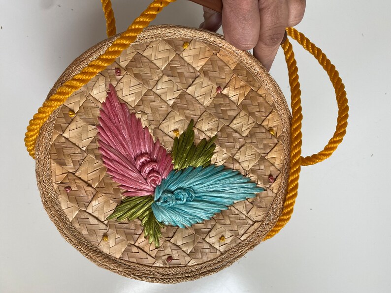 Op de afbeelding: Een ronde geweven strooien tas met een gele touwhandvat. De tas heeft een bloemenmotief met roze, groene en blauwe accenten.