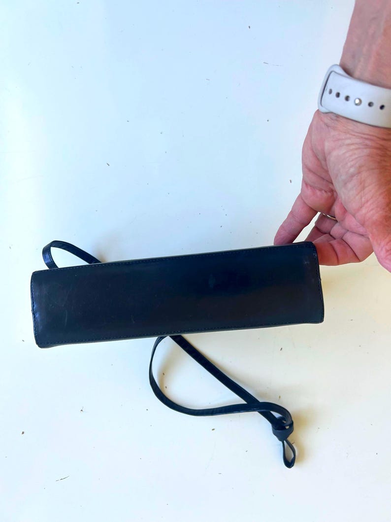 Pode incluir: Uma bolsa retangular preta com uma al&ccedil;a fina. A bolsa &eacute; feita de um material brilhante e tem um design simples e elegante. A al&ccedil;a &eacute; em la&ccedil;o e amarrada na extremidade.