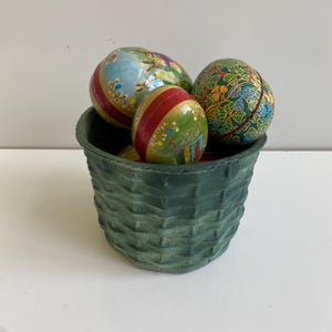 May include: A green woven basket filled with four decorative Easter eggs. The eggs are painted with various floral and geometric patterns in shades of green, red, yellow, blue, and white.