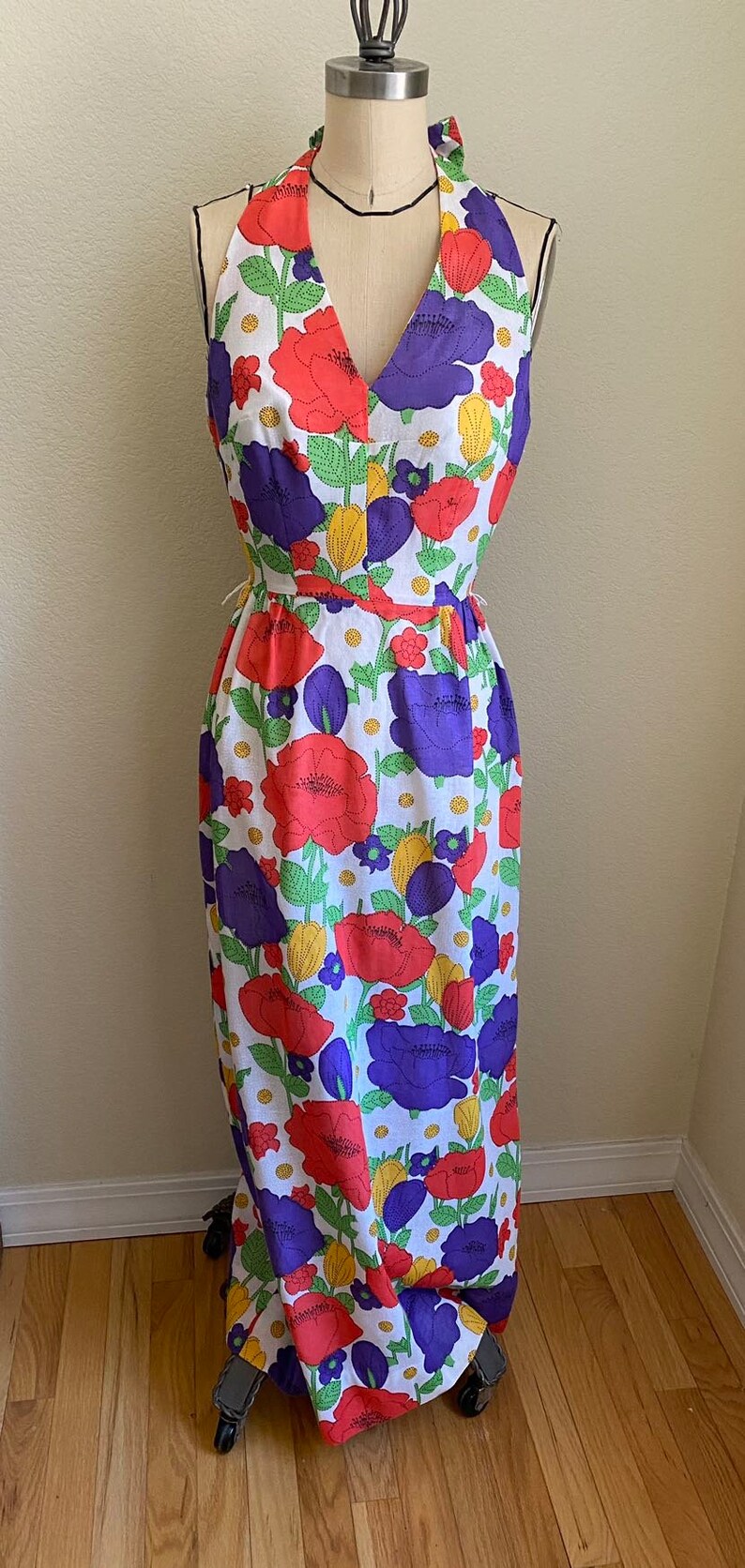 May include: A vintage halter-neck maxi dress with a vibrant floral pattern. The dress features large red and purple flowers with green leaves on a white background. The dress is displayed on a dress form.
