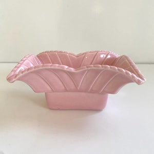May include: A light pink ceramic bowl with a square base and a scalloped, leaf-like rim. The bowl has a glossy finish and is likely vintage or retro in style. The bowl is empty and sits on a white surface.