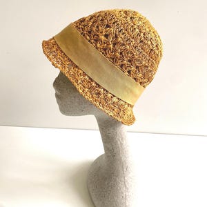 May include: A straw-colored cloche hat with a wide, light brown band. The hat is made of woven straw and sits on a mannequin head. The hat's brim curves slightly downward, and the band encircles the crown.