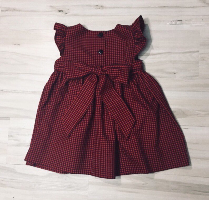 May include: A red and white gingham dress with a bow at the back. The dress has a sleeveless design and a ruffled neckline.