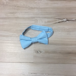 May include: A light blue bow tie made of fabric. The bow tie is on a light wood background.