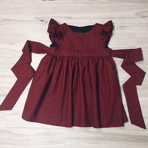 May include: Red and black gingham dress with ruffled sleeves and a black bow tie. The dress has a gathered skirt and a fitted bodice.