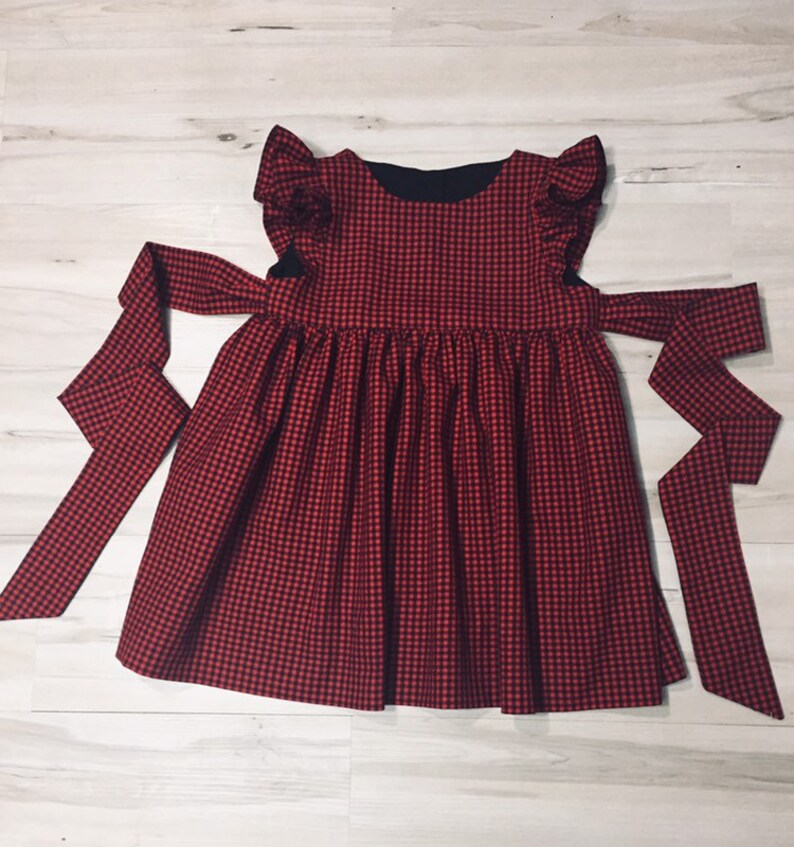 May include: Red and black gingham dress with ruffled sleeves and a black bow tie. The dress has a gathered skirt and a fitted bodice.