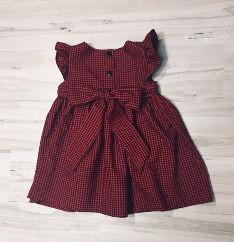 May include: A red and black gingham dress with a bow at the waist and ruffle sleeves.