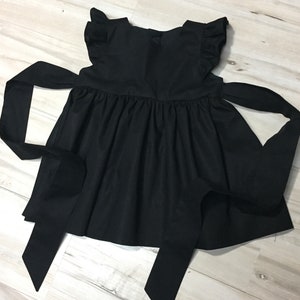 May include: A black sleeveless dress with a ruffled neckline and a bow at the back. The dress has a full skirt and two long ties that can be tied at the waist.