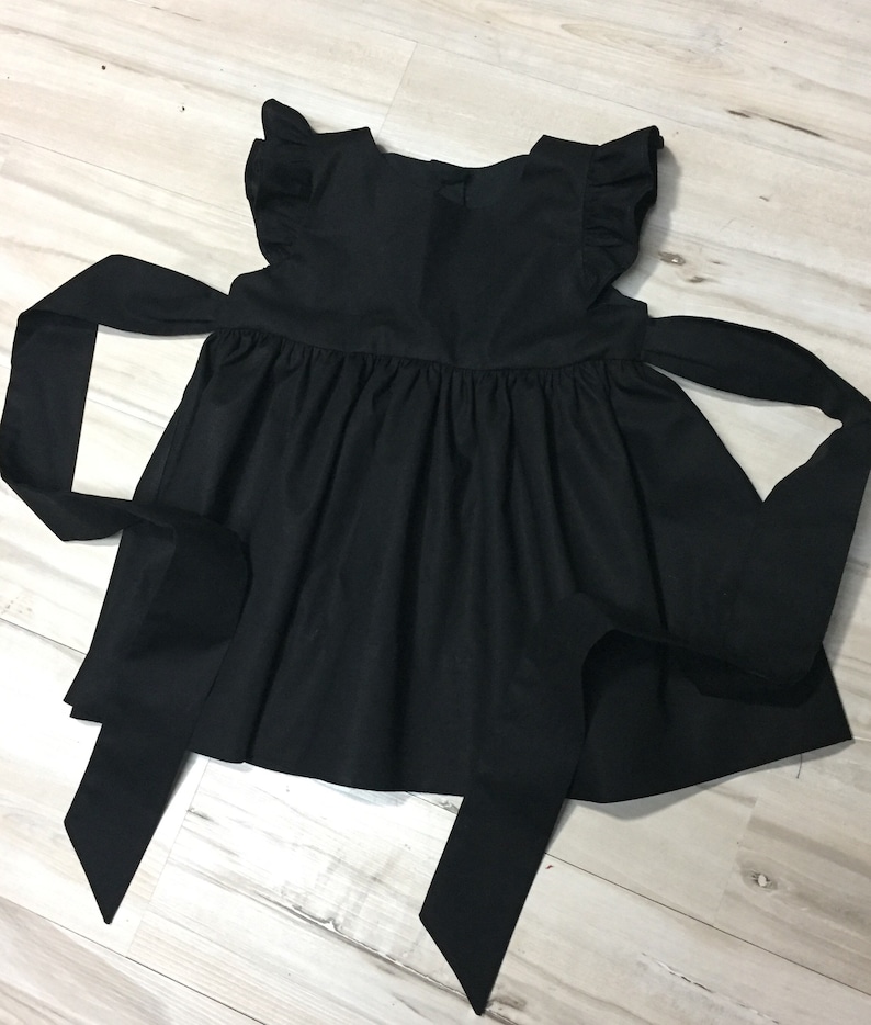May include: A black sleeveless dress with a ruffled neckline and a bow at the back. The dress has a full skirt and two long ties that can be tied at the waist.