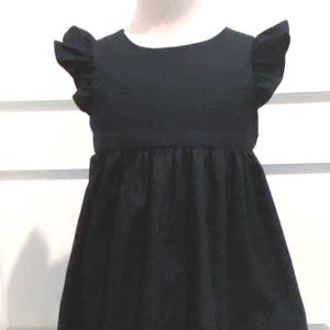 May include: A black toddler girl's dress with ruffled cap sleeves and a gathered waistline. The dress is made of a solid, dark fabric and has a simple, classic design. It is displayed on a mannequin against a white background.