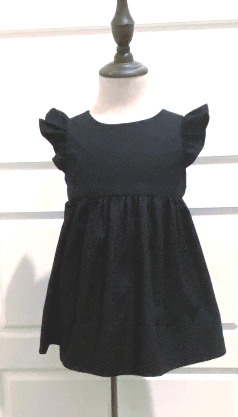 May include: A black toddler girl's dress with ruffled cap sleeves and a gathered waistline. The dress is made of a solid, dark fabric and has a simple, classic design. It is displayed on a mannequin against a white background.