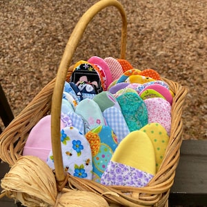 May include: A wicker basket filled with colorful, patterned fabric items. The items appear to be small, possibly baby hats, with various designs including floral, polka dots, and gingham. The basket has a woven handle and is tied with a raffia bow.