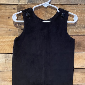 May include: Black corduroy overall romper with black buttons. The romper is hanging on a white hanger against a wood background.