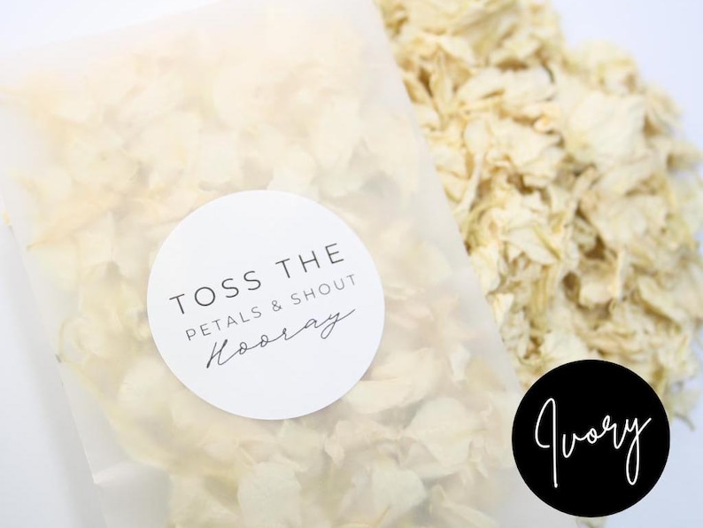 Puede incluir: Una bolsa de organza blanca llena de p&eacute;talos de flores de color marfil. La bolsa tiene una pegatina redonda blanca con el texto "Toss the Petals & Shout Hooray".