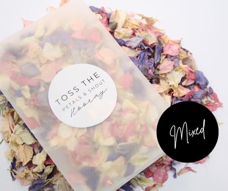 Puede incluir: Una bolsa de pl&aacute;stico transparente llena de una mezcla de p&eacute;talos de flores secas en tonos de rosa, blanco y morado. La bolsa tiene una pegatina circular blanca con el texto "Toss the Petals & Shout Hooray". Una pegatina circular negra con el texto "Mixed" est&aacute; en la mesa junto a la bolsa.