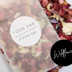 Puede incluir: Una bolsa de papel blanca llena de p&eacute;talos de flores rojos, rosas y blancos. La bolsa tiene una pegatina blanca que dice "Toss the petals & shout Hooray" y una pegatina negra que dice "Wildflower".