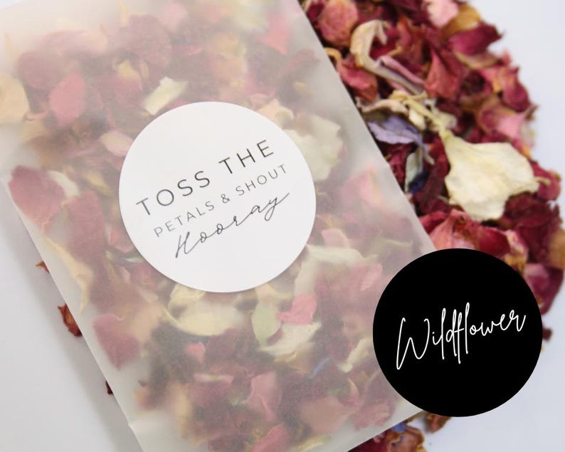 Puede incluir: Una bolsa de papel blanca llena de p&eacute;talos de flores rojos, rosas y blancos. La bolsa tiene una pegatina blanca que dice "Toss the petals & shout Hooray" y una pegatina negra que dice "Wildflower".