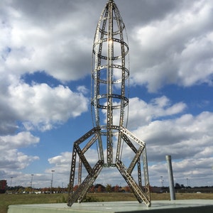 May include: A large, silver, metal rocket sculpture stands tall against a cloudy blue sky. The rocket is made of a latticework of metal bars and has a pointed top.