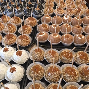 Op de afbeelding: Een assortiment van karamel appels met verschillende toppings, waaronder noten, sprinkles en kokosvlokken. De appels zijn gerangschikt in rijen op een zwart oppervlak.