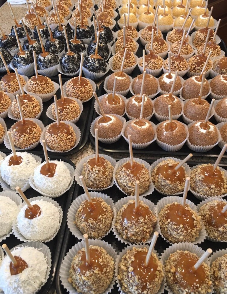 Op de afbeelding: Een assortiment van karamel appels met verschillende toppings, waaronder noten, sprinkles en kokosvlokken. De appels zijn gerangschikt in rijen op een zwart oppervlak.