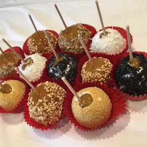Op de afbeelding: Een dozijn karamel appels met verschillende toppings, waaronder noten, kokosvlokken en chocoladesprinkels. De appels zijn gerangschikt in rode papieren bekers op een wit tafelkleed.
