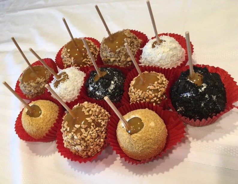 Op de afbeelding: Een dozijn karamel appels met verschillende toppings, waaronder noten, kokosvlokken en chocoladesprinkels. De appels zijn gerangschikt in rode papieren bekers op een wit tafelkleed.