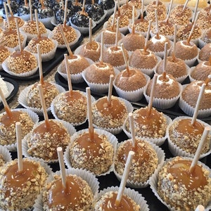 Op de afbeelding: Een close-up van een verscheidenheid aan karamel appels, sommige bedekt met noten, andere met chocolade en sprinkles. De appels zijn gerangschikt op een witte schaal met witte papieren voeringen.