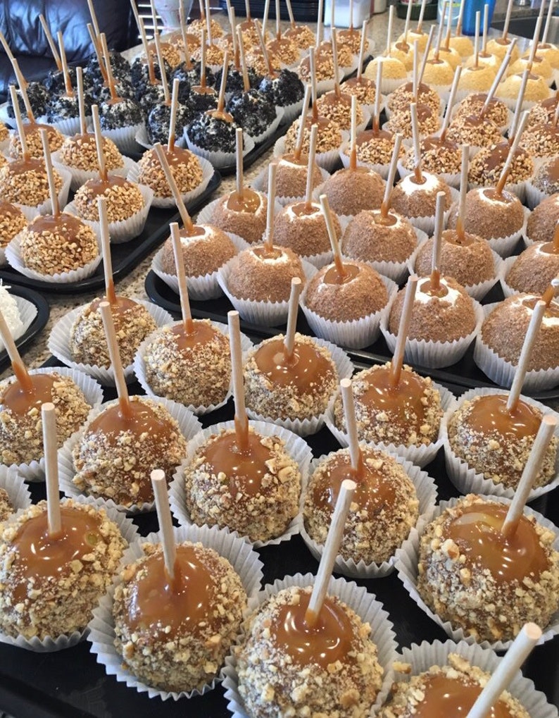 Op de afbeelding: Een close-up van een verscheidenheid aan karamel appels, sommige bedekt met noten, andere met chocolade en sprinkles. De appels zijn gerangschikt op een witte schaal met witte papieren voeringen.