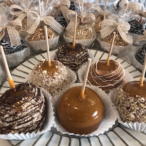 Op de afbeelding: Een schaal met karamel appels versierd met verschillende toppings, waaronder chocolade, noten en witte chocolade drizzle. De appels zijn gerangschikt op een witte en zilveren schaal.