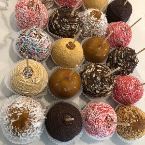Op de afbeelding: Assortiment van karamel appels versierd met verschillende toppings, waaronder sprinkels, chocolade, noten en graham cracker kruimels. De appels zijn gerangschikt in rijen op een wit oppervlak.
