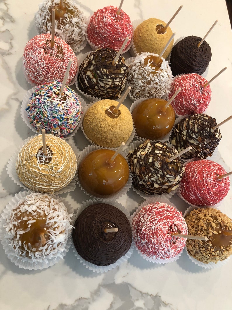 Op de afbeelding: Assortiment van karamel appels versierd met verschillende toppings, waaronder sprinkels, chocolade, noten en graham cracker kruimels. De appels zijn gerangschikt in rijen op een wit oppervlak.