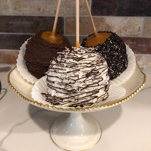 Op de afbeelding: Drie karamel appels op een witte voetstuk taartstandaard met een gouden rand. De appels zijn bedekt met chocolade, witte chocolade en kokosnoot, en chocoladesprinkels.