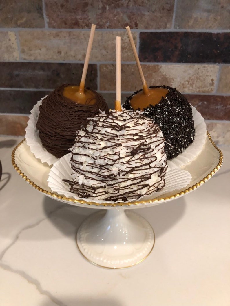 Op de afbeelding: Drie karamel appels op een witte voetstuk taartstandaard met een gouden rand. De appels zijn bedekt met chocolade, witte chocolade en kokosnoot, en chocoladesprinkels.