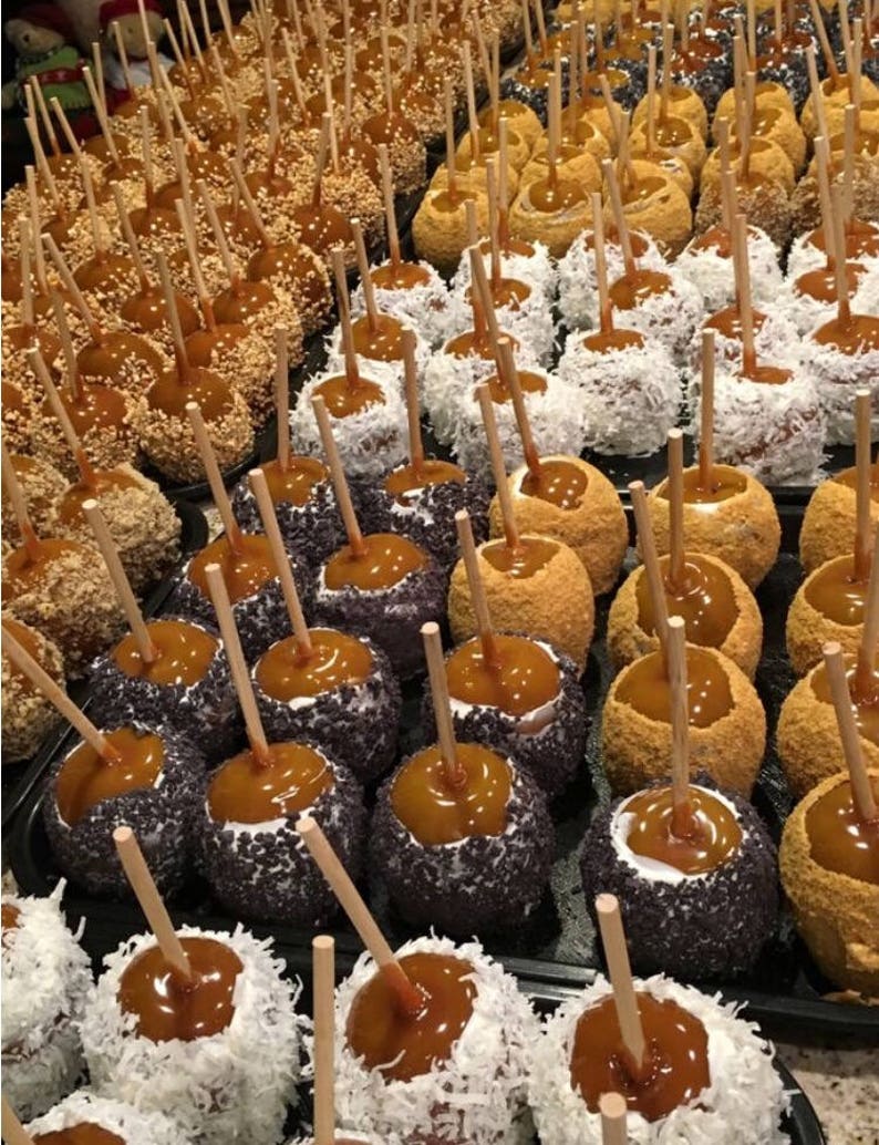 Op de afbeelding: Een assortiment karamel appels versierd met verschillende toppings, waaronder sprinkels, kokosvlokken en gemalen noten. De appels zijn gerangschikt op een zwarte schaal en klaar om te worden gegeten.
