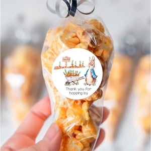 May include: A clear cellophane bag filled with orange popcorn, tied with a black ribbon. A round sticker features a watercolor illustration of a rabbit in a blue jacket, a wheelbarrow of carrots, and the text "Thank you for hopping by."