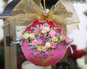 May include: A pink Christmas bauble decorated with a gold bow and ribbon flowers. The bauble is hanging from a gold string with a bell at the end.