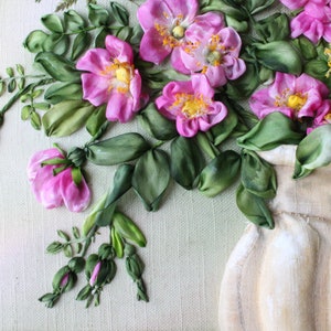 May include: A bouquet of pink silk ribbon roses in a cream-coloured vase. The roses are arranged in a cluster with green ribbon leaves. The background is a light green canvas.